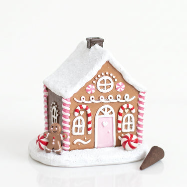 A small gingerbread house decoration with a white snow-dusted roof and base, pink door, red-and-white striped candy cane accents, and a tiny gingerbread man figure beside it on a white background.